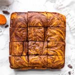 Pumpkin Swirl Brownies next to a glass of milk, a spoon of pumpkin puree, and chocolate chunks
