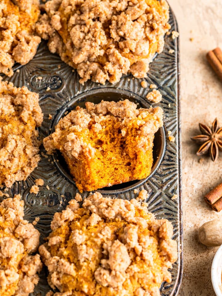 Pumpkin Crumb Cake Muffins in muffin pan surrounded by fall spices with one muffin missing a bite