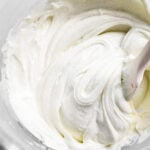 Close up of smooth cream cheese frosting and a silicone spatula inside a clear mixing bowl.