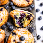 Close up of lemon blueberry muffin with a bite missing in muffin pan with blueberries and lemons surrounding the pan.