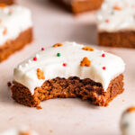 Close up of a bite missing from a gingerbread cookie bar with cream cheese frosting and holiday sprinkles on top.