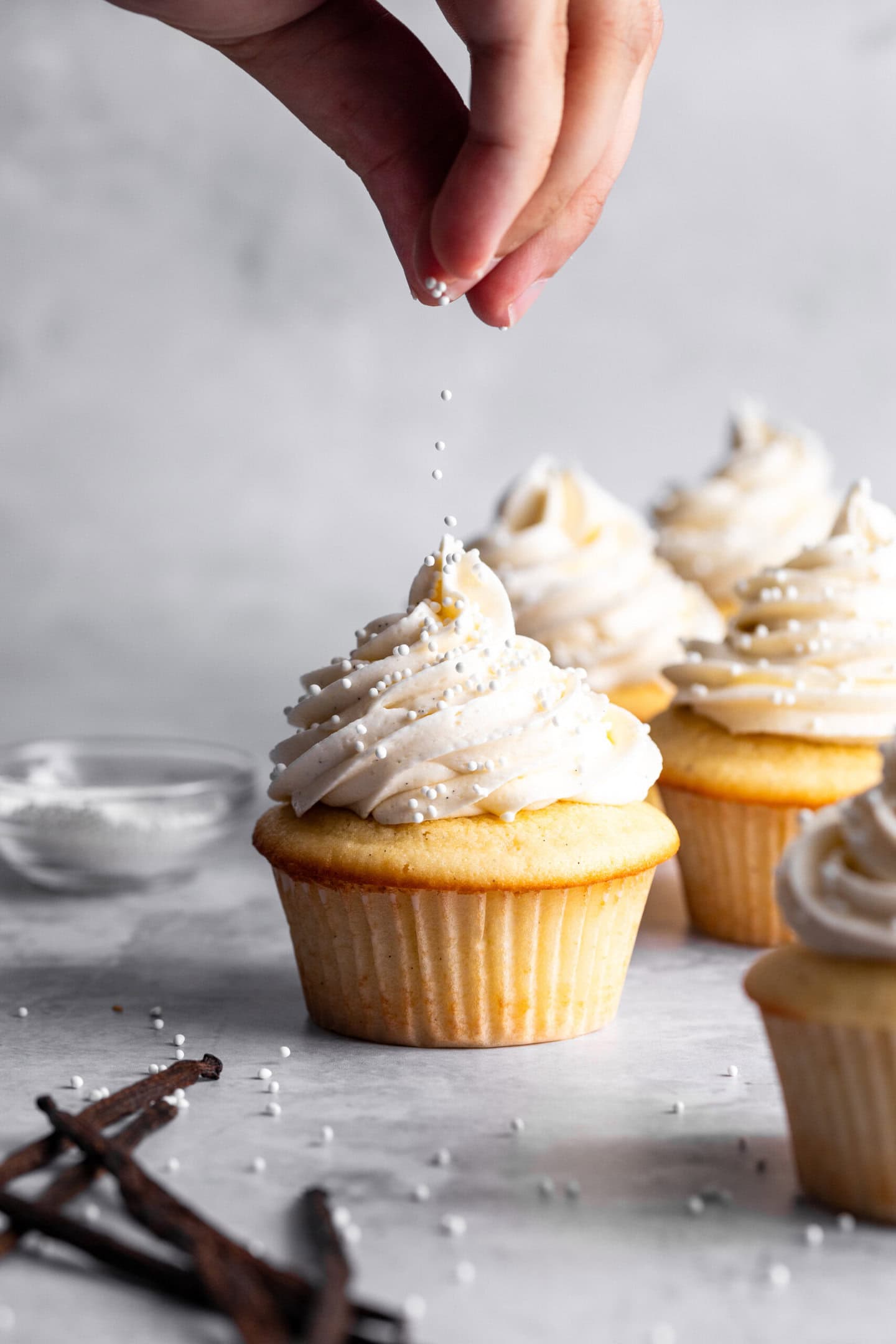 Sprinkles being dropped on top of vanilla cupcakes with vanilla frosting swirled on top.