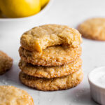 Lemon sugar cookies stacked next to a bowl of lemons and sparkling sugar. A bite is missing from the top cookie showing the texture of the inside.