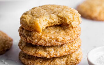 Lemon sugar cookies stacked next to a bowl of lemons and sparkling sugar. A bite is missing from the top cookie showing the texture of the inside.