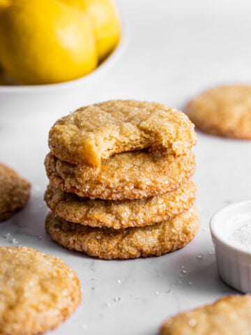 Lemon sugar cookies stacked next to a bowl of lemons and sparkling sugar. A bite is missing from the top cookie showing the texture of the inside.