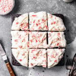 Slices of chocolate peppermint snack cake with peppermint cream cheese frosting swirled on top next to a small bowl of crushed candy canes, a glass of milk, forks, and an icing spatula.
