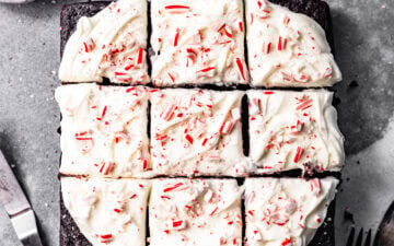Slices of chocolate peppermint snack cake with peppermint cream cheese frosting swirled on top next to a small bowl of crushed candy canes, a glass of milk, forks, and an icing spatula.