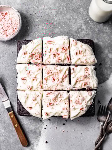 Slices of chocolate peppermint snack cake with peppermint cream cheese frosting swirled on top next to a small bowl of crushed candy canes, a glass of milk, forks, and an icing spatula.