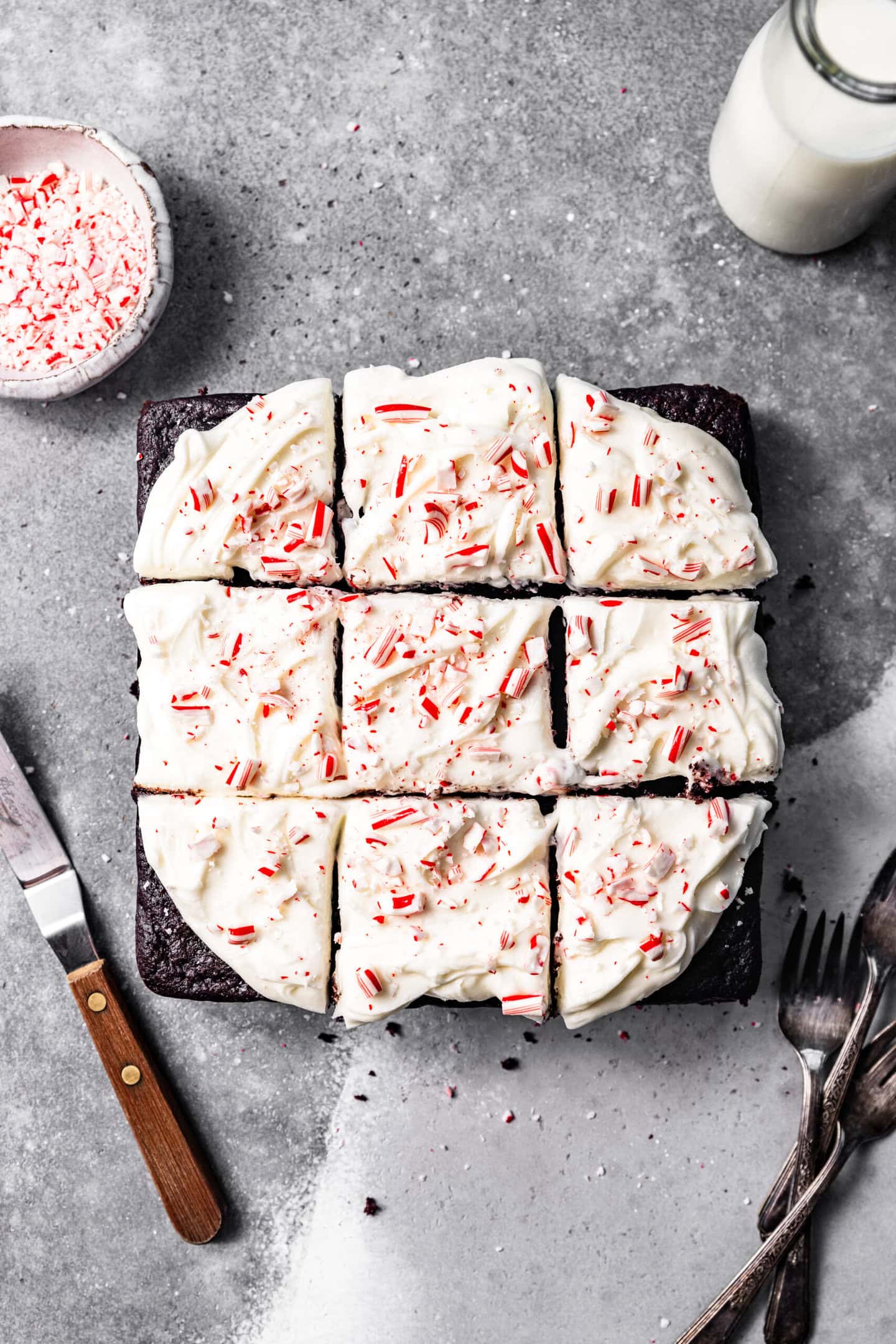 Slices of chocolate peppermint snack cake with peppermint cream cheese frosting swirled on top next to a small bowl of crushed candy canes, a glass of milk, forks, and an icing spatula.