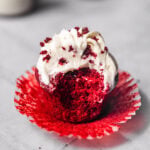 Close up of one red velvet cupcake with cream cheese frosting piped on top and a bite missing showing off the moist, fluffy interior.
