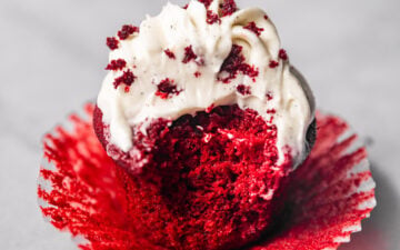 Close up of one red velvet cupcake with cream cheese frosting piped on top and a bite missing showing off the moist, fluffy interior.