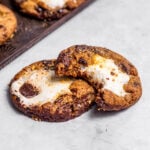 Marshmallow-stuffed s'mores cookies with a bite missing from one cookie and fresh baked cookies on a baking tray nearby.