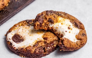Marshmallow-stuffed s'mores cookies with a bite missing from one cookie and fresh baked cookies on a baking tray nearby.