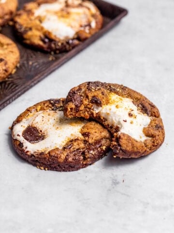 Marshmallow-stuffed s'mores cookies with a bite missing from one cookie and fresh baked cookies on a baking tray nearby.