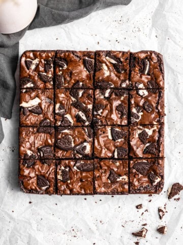 Oreo Brownies on top of parchment paper with pieces of Oreo cookies and a glass of milk and napkin nearby.