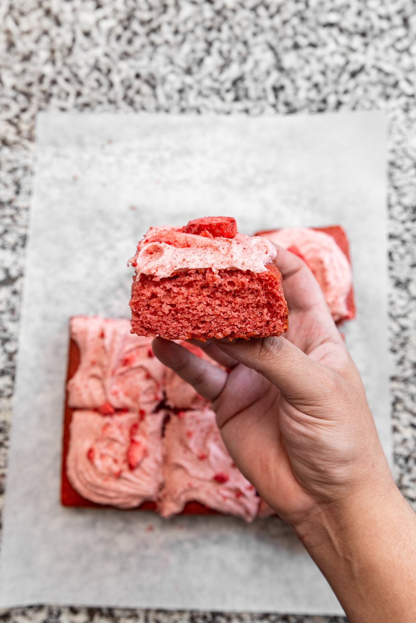 A hand holding up a slice of strawberry cake showing off it's moist, fluffy texture and creamy frosting on top.