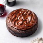 A Single-Layer Red Wine Chocolate Cake with red wine chocolate ganache on top surrounded by plates, forks, and a bottle of red wine with a filled wine glass.