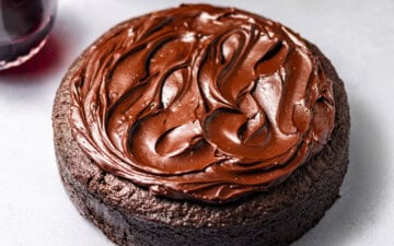A Single-Layer Red Wine Chocolate Cake with red wine chocolate ganache on top surrounded by plates, forks, and a bottle of red wine with a filled wine glass.