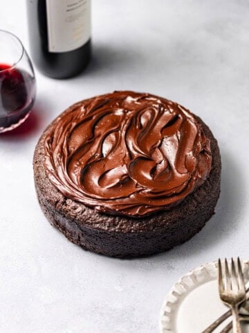 A Single-Layer Red Wine Chocolate Cake with red wine chocolate ganache on top surrounded by plates, forks, and a bottle of red wine with a filled wine glass.