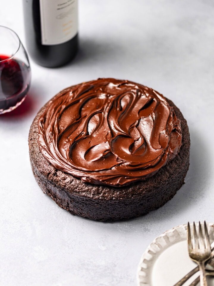 A Single-Layer Red Wine Chocolate Cake with red wine chocolate ganache on top surrounded by plates, forks, and a bottle of red wine with a filled wine glass.