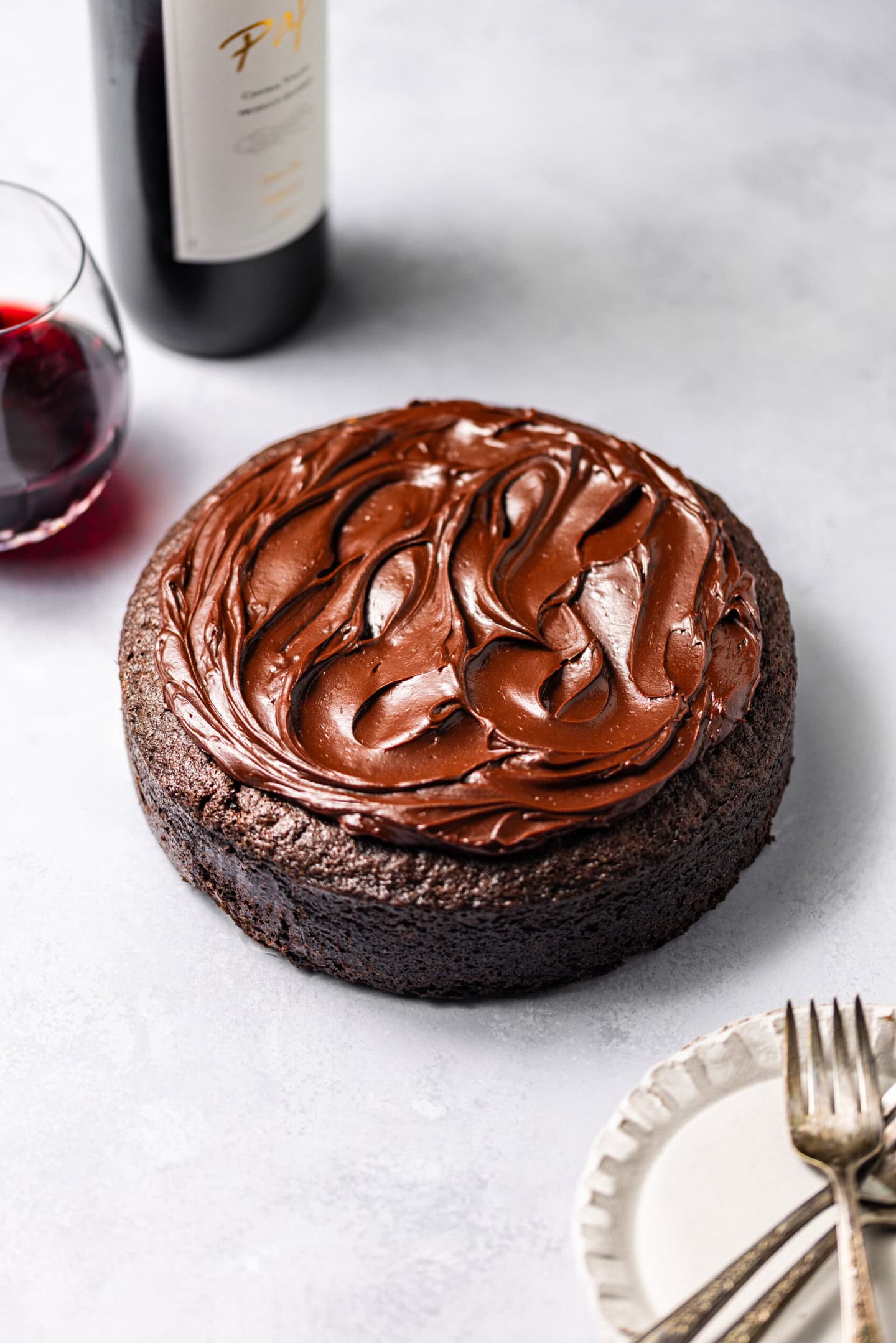 A Single-Layer Red Wine Chocolate Cake with red wine chocolate ganache on top surrounded by plates, forks, and a bottle of red wine with a filled wine glass.