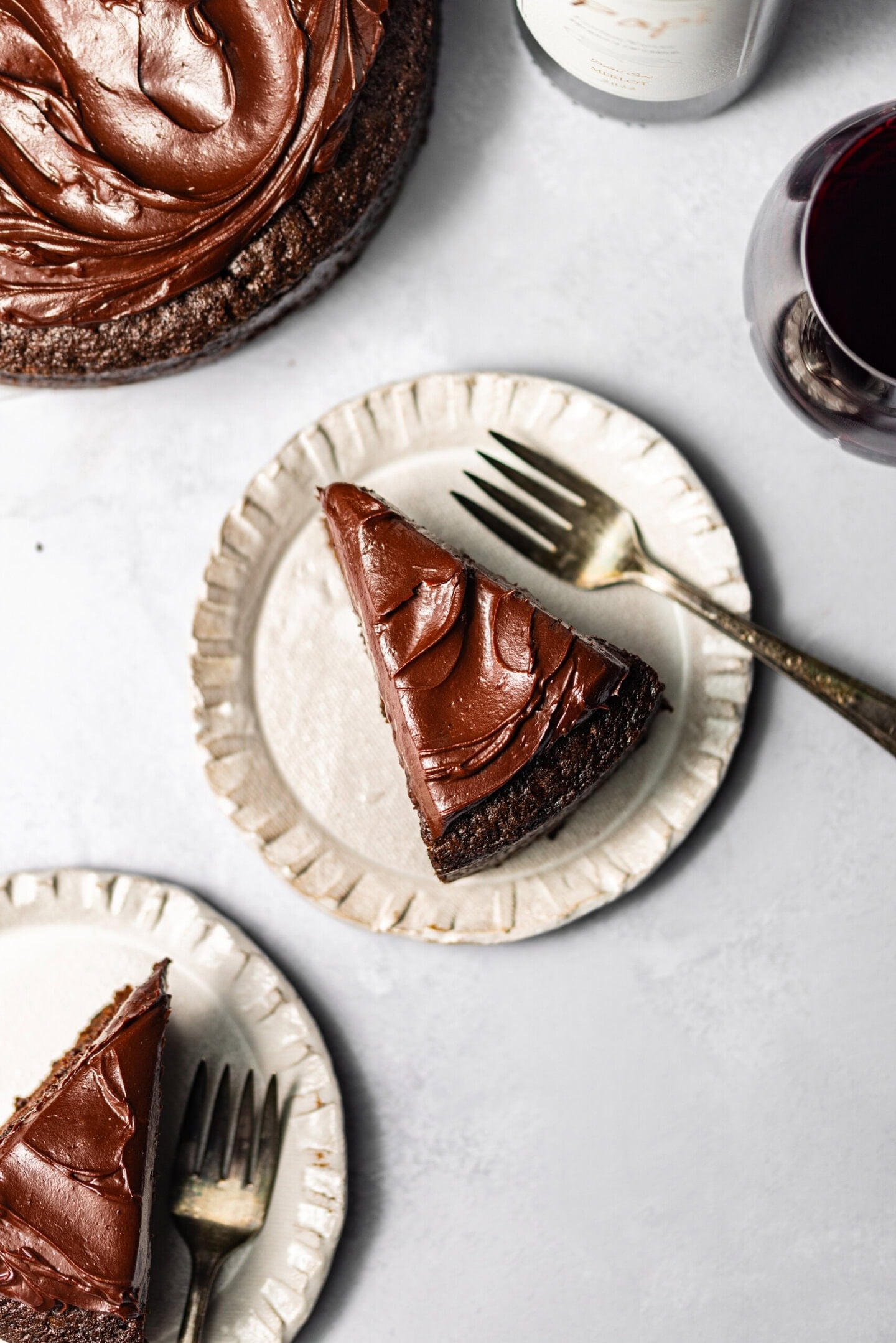 Slices of red wine chocolate cake with chocolate ganache on top on plates with forks next to each slice and a wine glass nearby.