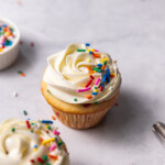 Two funfetti cupcakes with vanilla buttercream frosting and rainbow sprinkles on top sitting next to a small pinch pot of sprinkles and a piping bag filled with frosting.