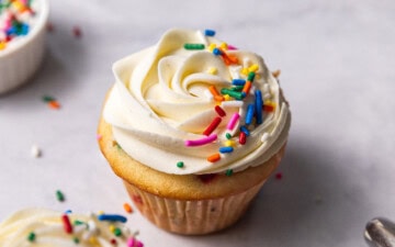 Two funfetti cupcakes with vanilla buttercream frosting and rainbow sprinkles on top sitting next to a small pinch pot of sprinkles and a piping bag filled with frosting.
