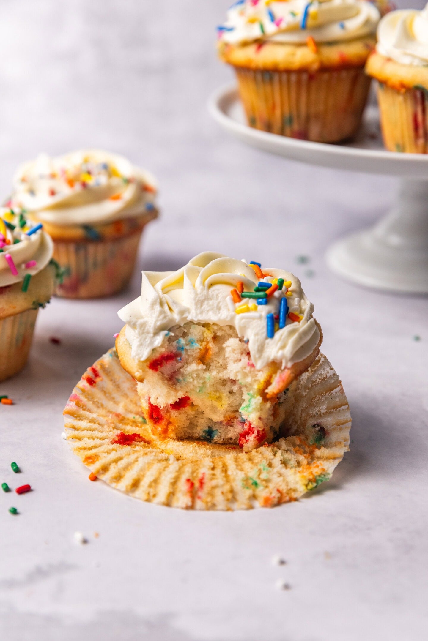 A bite taken out of a cupcake showing the moist, soft and fluffy interior with pops of color from the rainbow sprinkles. Other funfetti cupcakes are sitting in the background on top of a cake stand.